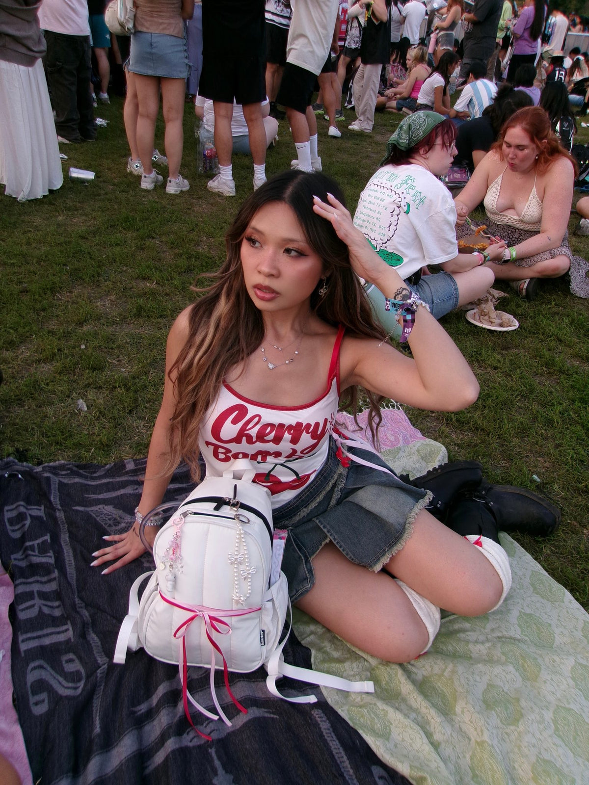 Woman sitting on grass with a white backpack, surrounded by people in a casual outdoor setting.
