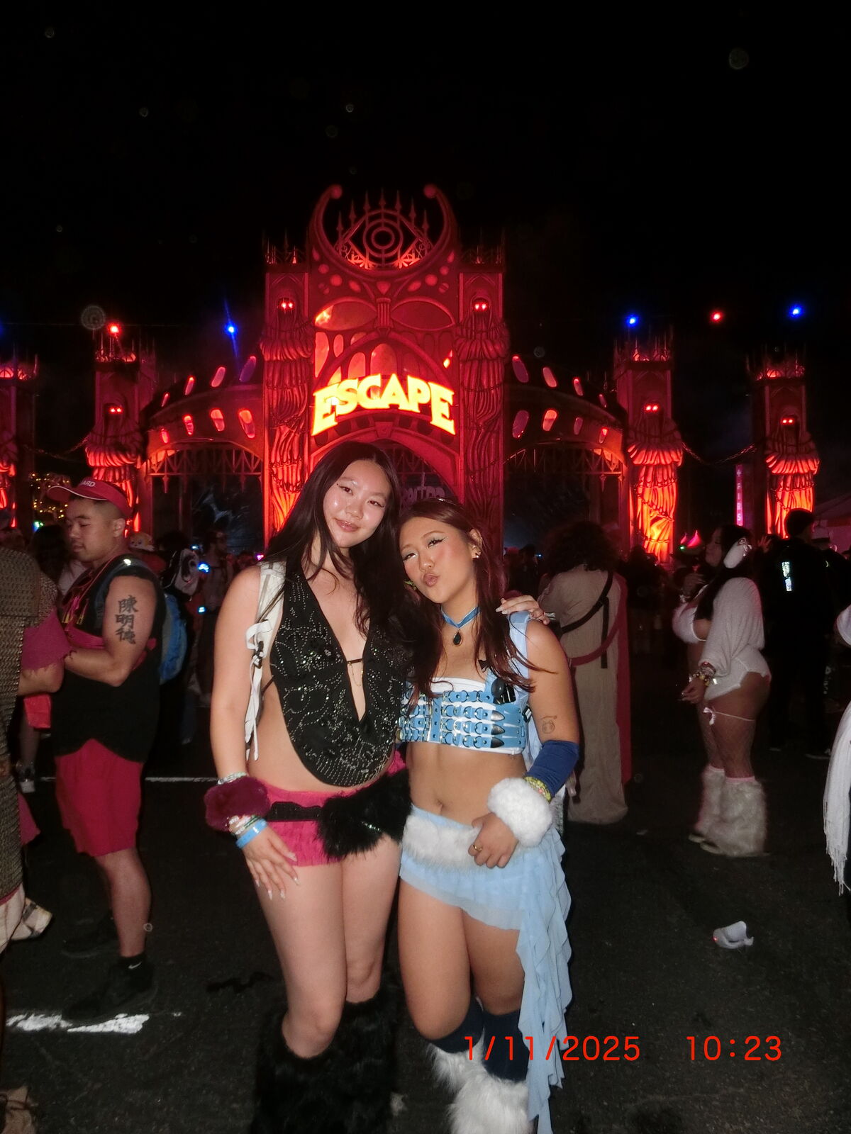 Two women posing in costume outfits in front of a brightly lit red 'ESCAPE' themed arch at night, with other people and a dark festival environment in the background.